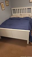 Photo of full size white wooden bed frame with slatted headboard and footboard, mattress present with bedding including pillows and navy blue comforter.