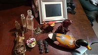 Photo showing all 13 pieces on a wooden table, including glass bottles, ceramic flowers, wood African carvings, duck decoy, French Bulldog figure, fisherman statue, cardinals, and electronic picture frame.