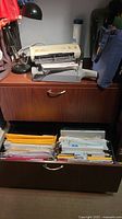 Metal two-drawer filing cabinet with wood grain finish, drawers open revealing paper files organized in hanging folders. Top surface holds an old typewriter and a desk lamp.