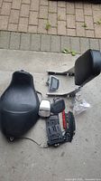 Photo showing the collection of Harley Davidson motorcycle parts including seat, sissy bar, side license plate holder, foot pegs, mirror, and small hardware pieces.