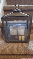 Medium metal and glass hurricane lantern with two flameless flickering pillar candles in original box displayed on carpeted stairs.