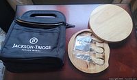 Front view of black Jackson-Triggs insulated wine carrier bag next to round wooden cheese board with metal utensils