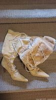 Beige knee-high boots laid flat showing laces and fur lining inside, pointed toe and high heel visible.