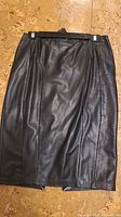 Front view of a black leather skirt hanging on a hanger, showing the smooth leather and vertical stitching details.