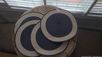 Four round woven placemats stacked in black center with beige border on table near window.