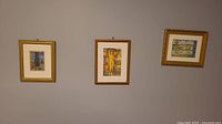 Three framed art prints hanging on wall in a row, two smaller and one medium size, varying wooden and gold colored frames.