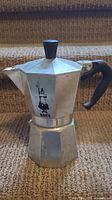 Silver aluminum stovetop espresso maker with black handle and knob, octagonal shape, minor scratches, and a logo of a man with a pipe.