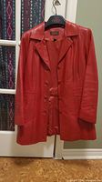 Front view of red leather Danier coat hanging on a hanger in front of a door, showing button closure and classic collar.
