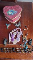 Heart-shaped jewelry box on wooden surface surrounded by assorted costume jewelry including earrings, bracelets, and brooches in mixed colors and materials