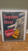Photo of vintage Kodak Film advertisement poster framed in metal, showing multicolor text and images with some corner wear and creasing.