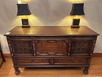 Front view of carved oak sideboard with two table lamps, showing three drawers and two carved floral panel doors on ball-and-claw feet