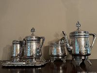 Full view of the silver plated tea set including teapot, coffee pot, creamer, sugar bowl, and tray showing matching embossed floral and geometric designs and decorative handles and feet.