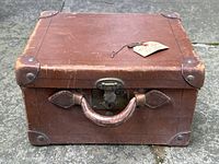 Front view of antique brown leather hat box showing leather handle, brass lock, corner reinforcement, and attached key tag.