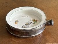 Side angle photo showing the ceramic bowl within the silver plated porringer and its handle, capturing the dented and worn condition of the silver plating.