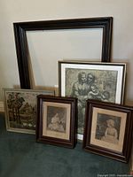 Five framed items displayed: one large empty black frame, three framed black and white prints with human figures, and a framed needlepoint landscape.