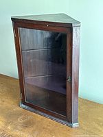 Front angled view of the antique corner cabinet showing glass door, triangular shape, and metal latch.