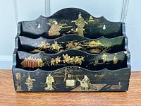 Front view of antique Japanese black lacquerware letter holder showing detailed gold and colored hand-painted figures and landscapes on three compartments and base.