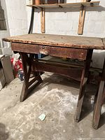 Front angle view of antique wooden workbench showing thick wooden top, drawer, and trestle legs