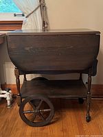 Side view of the walnut tea cart showing wooden wheel, turned leg, drop-leaf side and lower shelf
