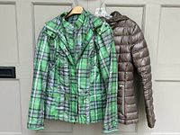 Green plaid rain jacket and taupe Calvin Klein down filled jacket hung on door, showing full fronts.