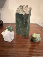 Three sculptures displayed: a large green soapstone block with raw edge in center, a green soapstone seal sculpture, and a green soapstone bear on a clear rock crystal base with a fish figure
