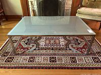 Clear front view of a rectangular coffee table with frosted glass top and steel frame placed on a patterned rug in a living room setting.