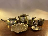 Similar angle photo of vintage brass kettles, decorative dish, candle holder, pitcher, bowls, and mold-like container on a wood surface.
