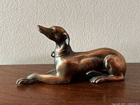 Copper greyhound figurine shown from left side, displaying smooth tarnished copper surface, detailed head and collar, and missing front paw.
