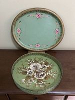 Both metal toleware trays shown resting on wooden furniture, displaying floral patterns and shapes