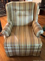 Front view of vintage armchair showing striped fabric upholstery, tufted back with buttons, rolled arms, and skirted bottom. Visible staining and wear on fabric.