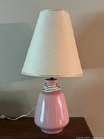 Full view of the vintage pink ceramic table lamp with beige tapered fabric lampshade against a light wall background.