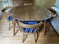 Oval extendable wooden dining table with four matching wooden spindle-back chairs featuring blue cushions, placed on wood flooring in a dining room.