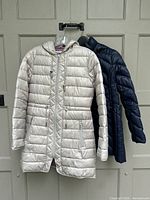 Two lightweight down jackets hanging together on a hanger against a door, one taupe and one navy blue, showing full length and front views.