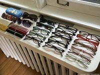 Wide view of 18 pairs of women's reading glasses and sunglasses arranged on a white surface, showing variety of styles, colors, and frame shapes.
