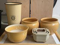 Full view of all seven garden planters on surface showing various sizes, colors and styles including the tall yellow enamel vase with printed logo and six ceramic planters/bowls.