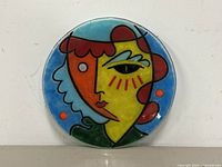 Full view of the circular glass plate showing the abstract woman's face artwork in vibrant colors and black outlines.