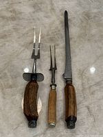Front view of the three antique meat carving tools laid out on a marble surface, showing the wooden handles and metal prongs/blade