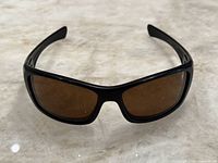 Front view of men's black framed Oakley sunglasses with brown lenses on marble surface.