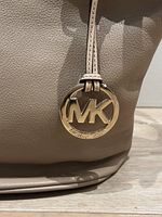 Close-up of gold-tone MK logo charm on taupe leather bag showing brand detail and leather texture