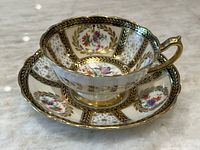 Teacup and saucer displayed on a marble surface showing hand painted floral patterns, gold accents, and black trimming from a front angle.