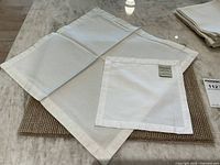 Photo showing four white Pottery Barn cocktail napkins displayed on a gold woven placemat on a marble table.