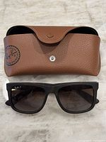 Brown textured leather Ray-Ban sunglasses case with snap button closure and logo, with Ray-Ban polarized sunglasses placed beneath it on a light surface.