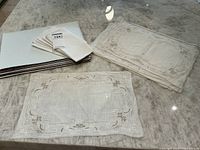 Set of white linen placemats stacked with some laid flat showing detailed embroidery and cutwork design patterns along edges and center areas.