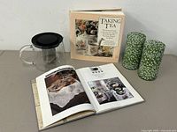 Glass teapot with black lid and strainer inside, two books standing upright, and two green floral canisters placed beside them.