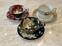 Photo showing all three teacup and saucer sets on a marble surface: Epiag white cup with gold and grey trim, Chugai black with cream floral, and Wako red with cabbage rose floral.