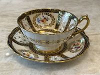Full image of teacup and saucer showing floral designs, gold accents, black trim, and overall condition