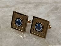 Pair of gold-tone metal cufflinks with blue enamel Toronto Maple Leafs logo and crossed hockey sticks on a textured square background.