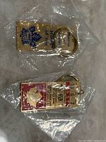 Two gold-toned metal Toronto Maple Leafs/Maple Leaf Gardens keychains in original plastic packaging, laid side by side, showing the front and back of the ticket keychain and the rectangular blue text keychain.