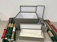 Photo showing the three grey gift boxes with lids and multiple rolls of wrapping paper in green, red, and cream colors bundled with rubber bands.