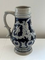 Front view of stoneware pitcher showing raised cherub designs and ornate detailing with dark blue and gray glaze.